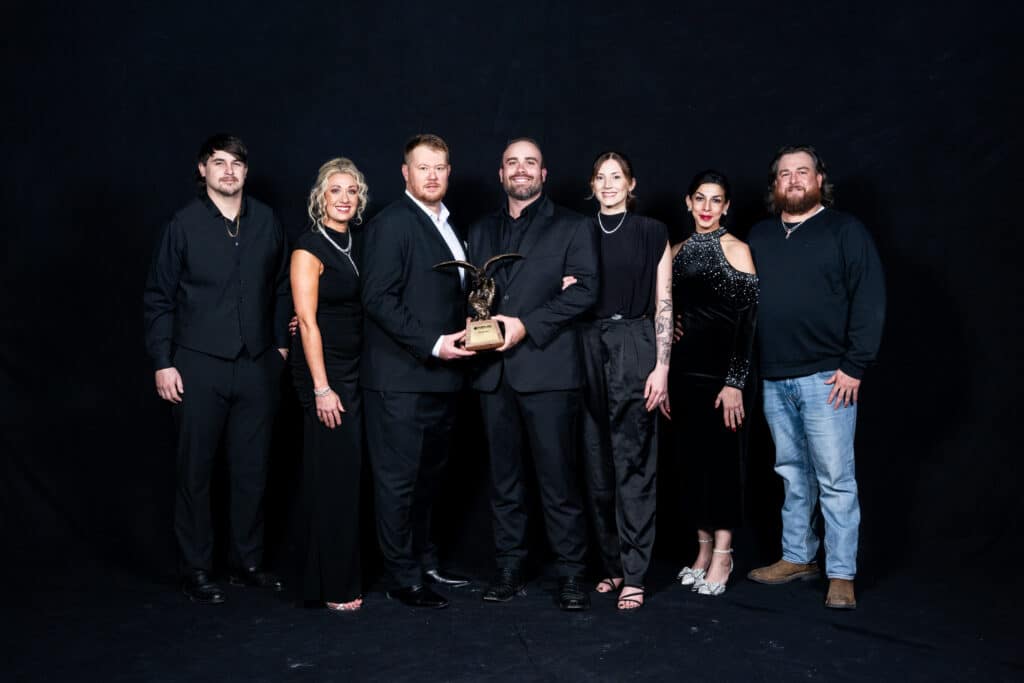 Seven people in formal and semi-formal attire stand together against a black background, with one person holding a trophy at the center.